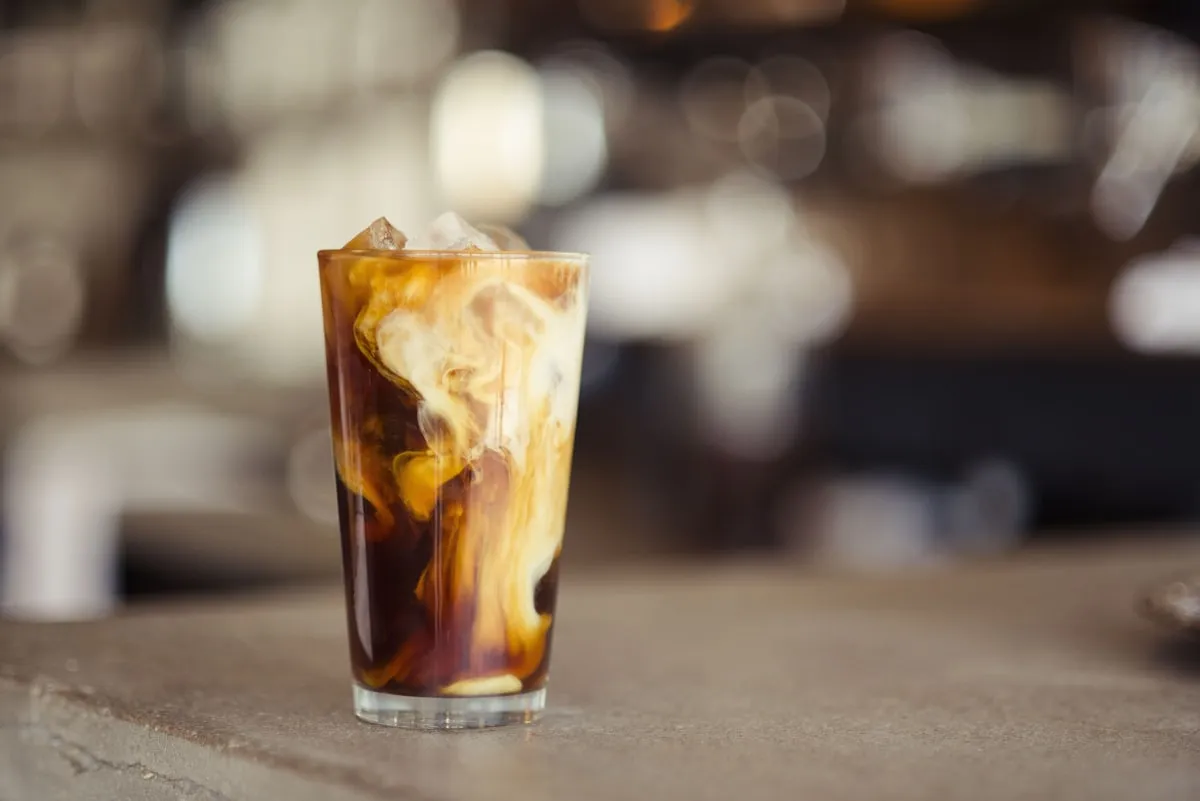 An iced coffee with cream swirling through it on a warm-toned counter with soft bokeh lighting in the background